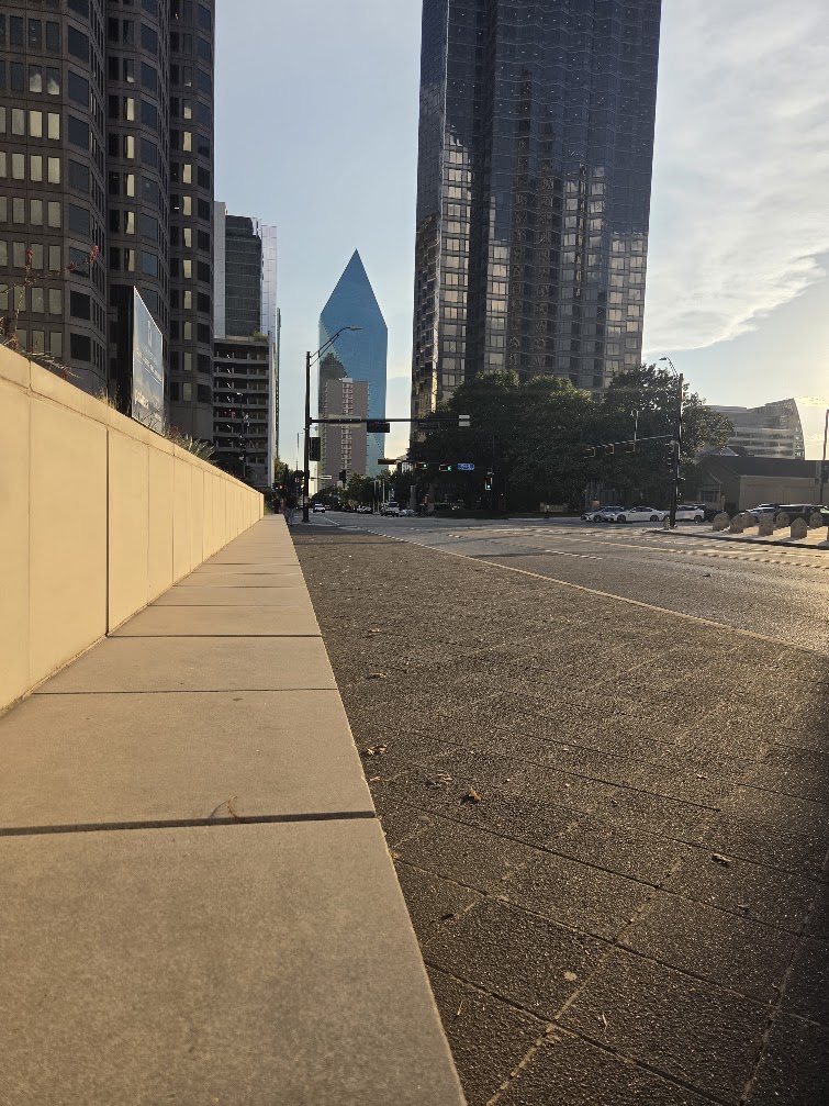 Downtown Dallas street with high-rise buildings and a blue glass tower in the distance, photographed in warm afternoon light.